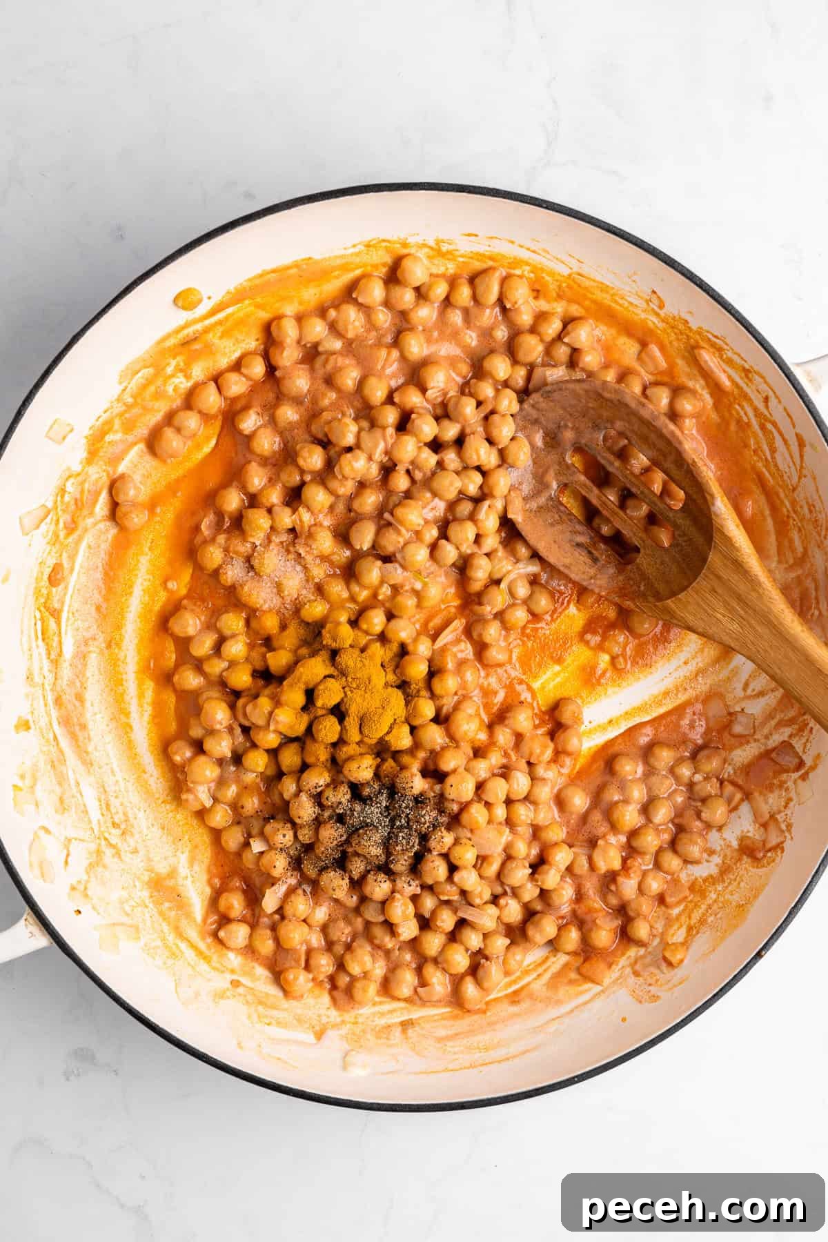 A skillet filled with garbanzo beans in a rich sauce, stirred with a wooden spoon. Seasonings including black pepper are visible on top. The background is a white marbled surface.