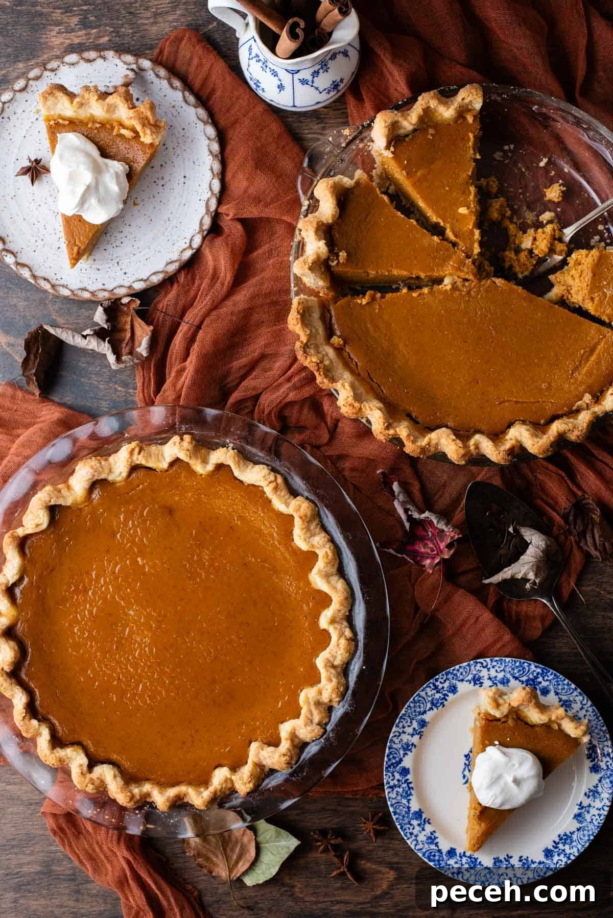 Classic Homemade Pumpkin Pie: Simple Recipe for Perfect Fall Dessert 2 an overhead view of a tablescape with two pumpkin pies, one whole and one sliced, sitting on top of a dark orange cloth on a wood surface and surrounded by two small plates with pumpkin pie slices topped with whipped cream, fall leaves and a dish of cinnamon sticks