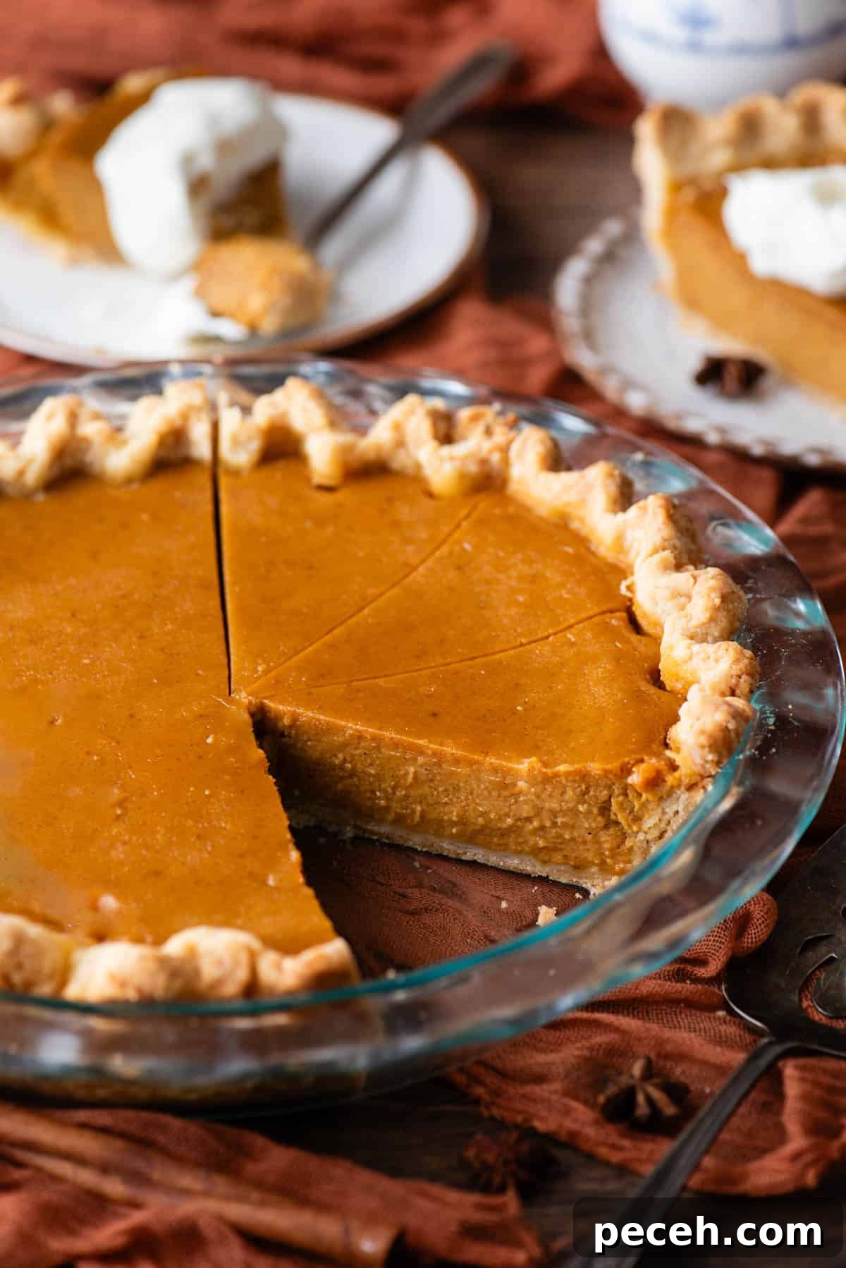 Classic Homemade Pumpkin Pie: Simple Recipe for Perfect Fall Dessert 11 pumpkin pie in a glass pie dish on a wood surface with a dark orange cloth under it, sliced with one slice missing and two plates in the background with pumpkin pie on them