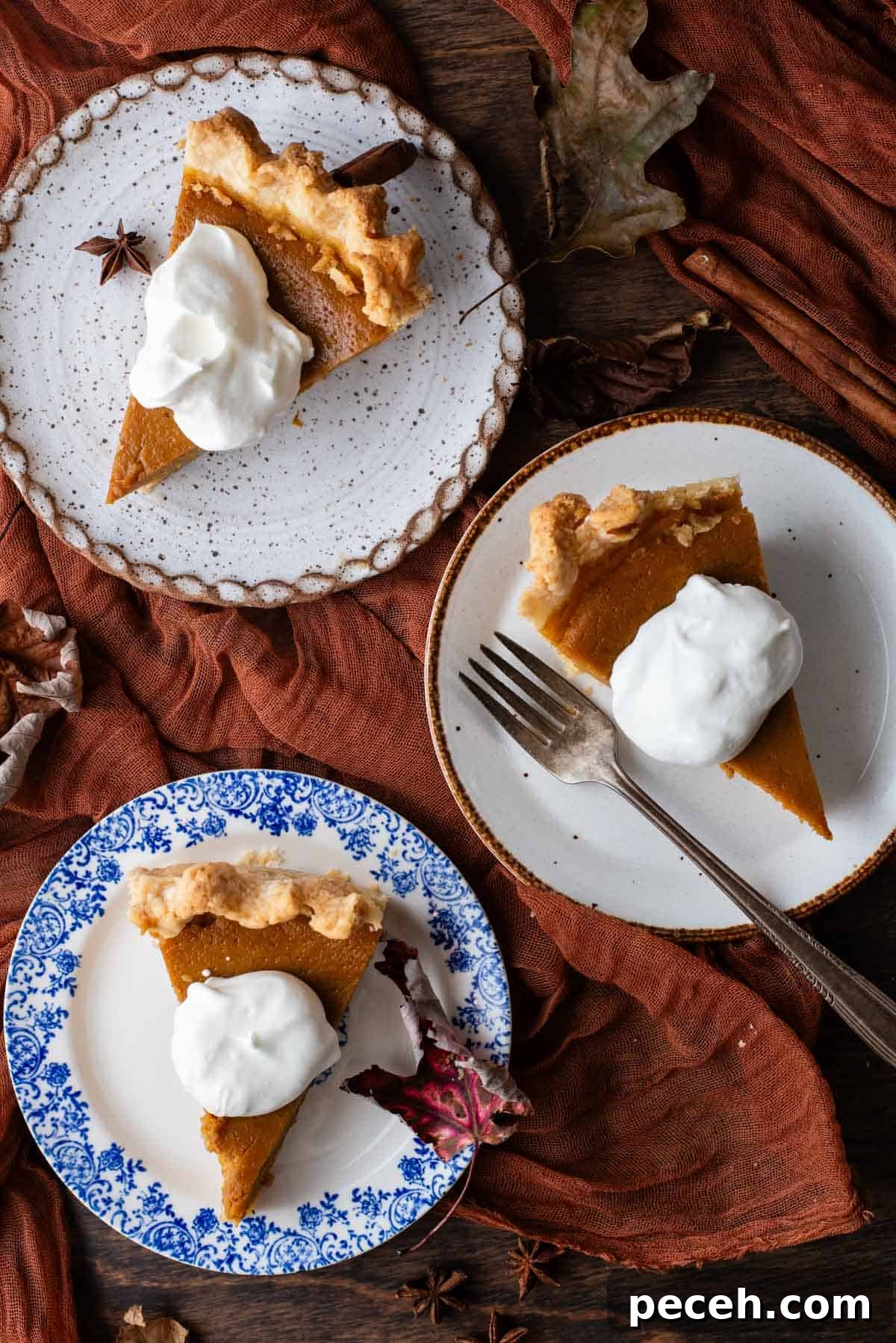 Classic Homemade Pumpkin Pie: Simple Recipe for Perfect Fall Dessert 14 three small plates sitting on a wood surface with a dark orange cloth and fall leaves scattered around, with slices of pumpkin pie on them topped with a dollop of whipped cream, and a fork on the middle plate