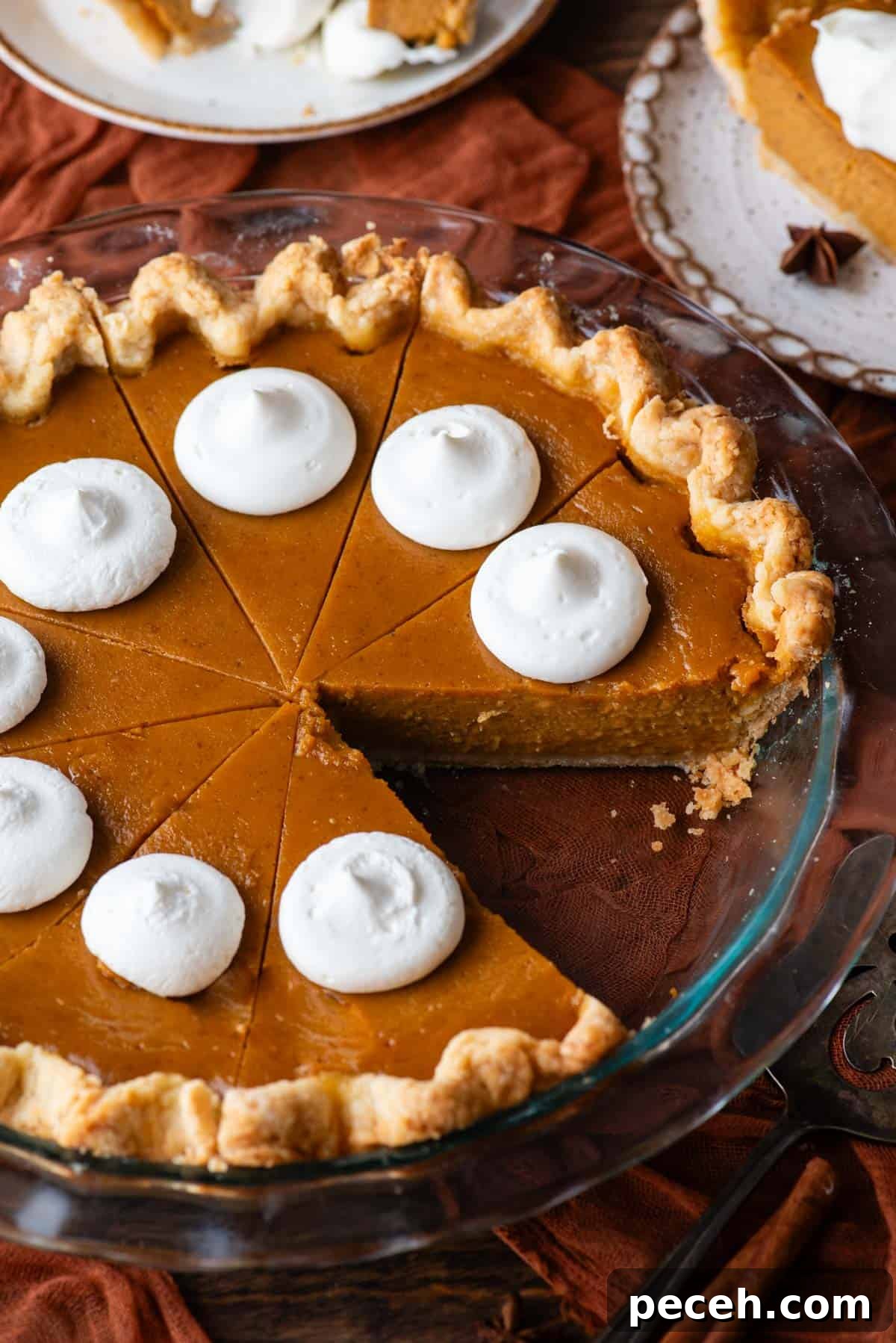 Classic Homemade Pumpkin Pie: Simple Recipe for Perfect Fall Dessert 4 a pumpkin pie in a glass pie dish with dollops of whipped cream on top, with one slice missing, sitting on top of a dark orange cloth on a wood surface with two plates with pumpkin pie on them beside it
