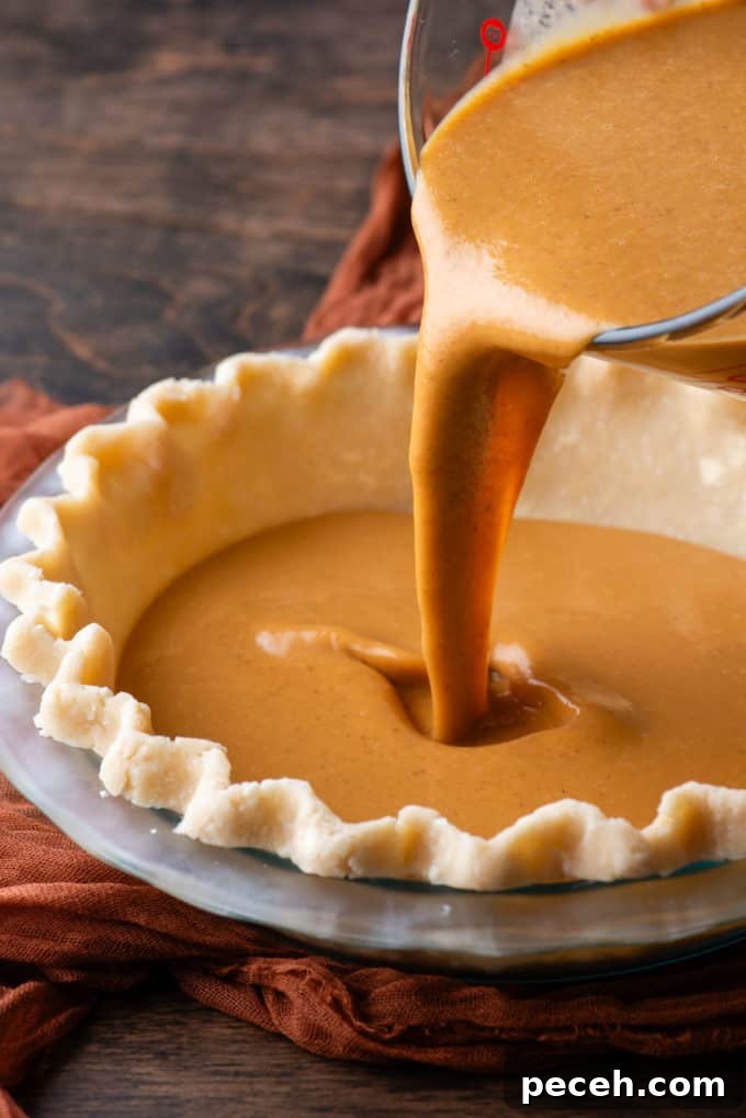 Classic Homemade Pumpkin Pie: Simple Recipe for Perfect Fall Dessert 9 pumpkin pie filling being poured into a glass pie dish with pie crust in it, which is sitting on top of a dark orange cloth on a wood surface