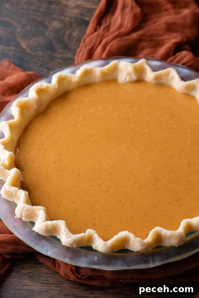 Classic Homemade Pumpkin Pie: Simple Recipe for Perfect Fall Dessert 10 an uncooked pumpin pie in a glass pie dish sitting on a dark orange cloth on a wood surface