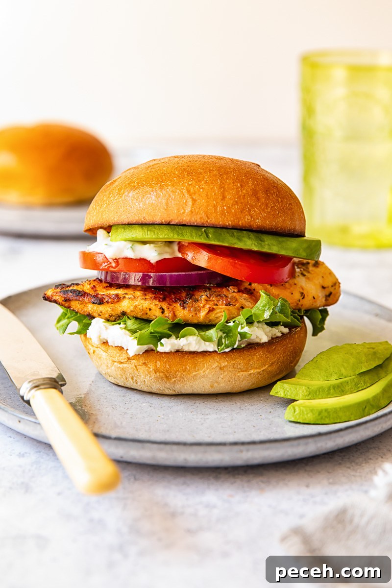 Greek Chicken Burger with Feta and Tzatziki Recipe 5 chicken burger on a grey plate, avocado slices, a butter knife, a green glass in the background.