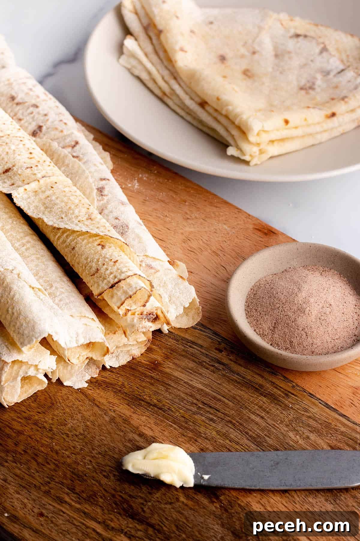 Authentic Norwegian Potato Lefse Recipe for Soft, Sweet Flatbread 2 A wooden board with rolled up lefse, a knife with butter, and a bowl with cinnamon sugar.