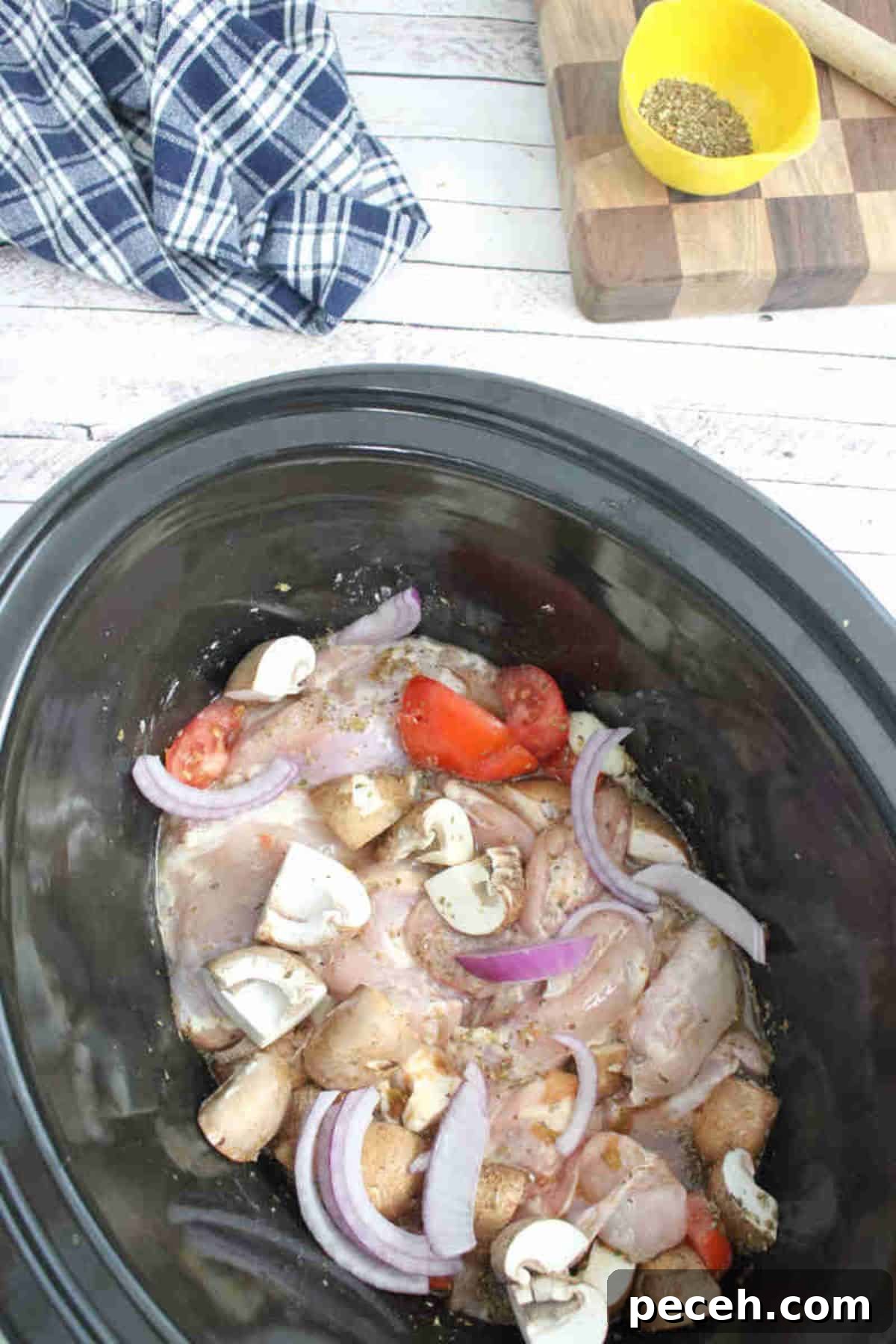 Raw chicken in a slow cooker with mushrooms, tomatoes and broth topped with red onions.
