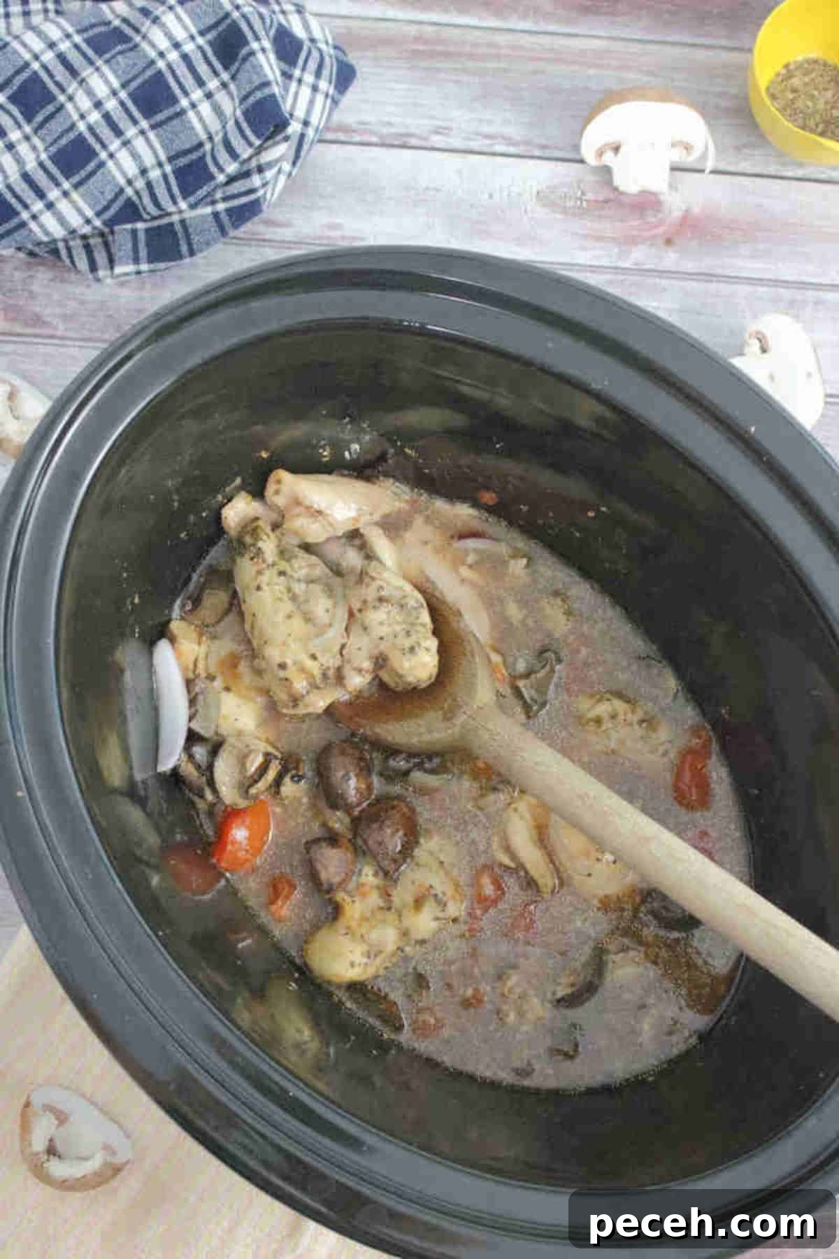 Wooden spoon in a slow cooker with cooked chicken thighs, tomatoes, and mushrooms.