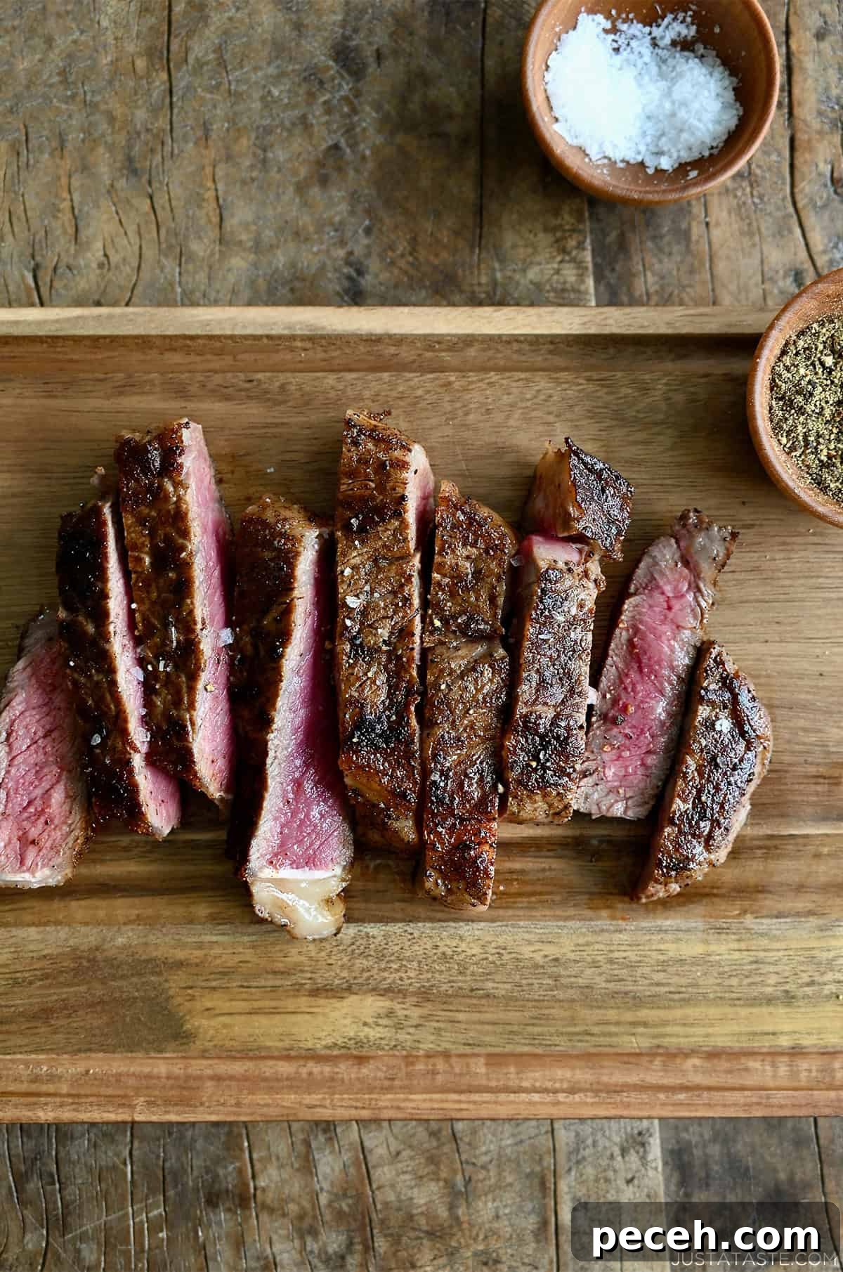 Sliced pan-seared steak on a wooden cutting board with small bowls containing salt and pepper nearby.