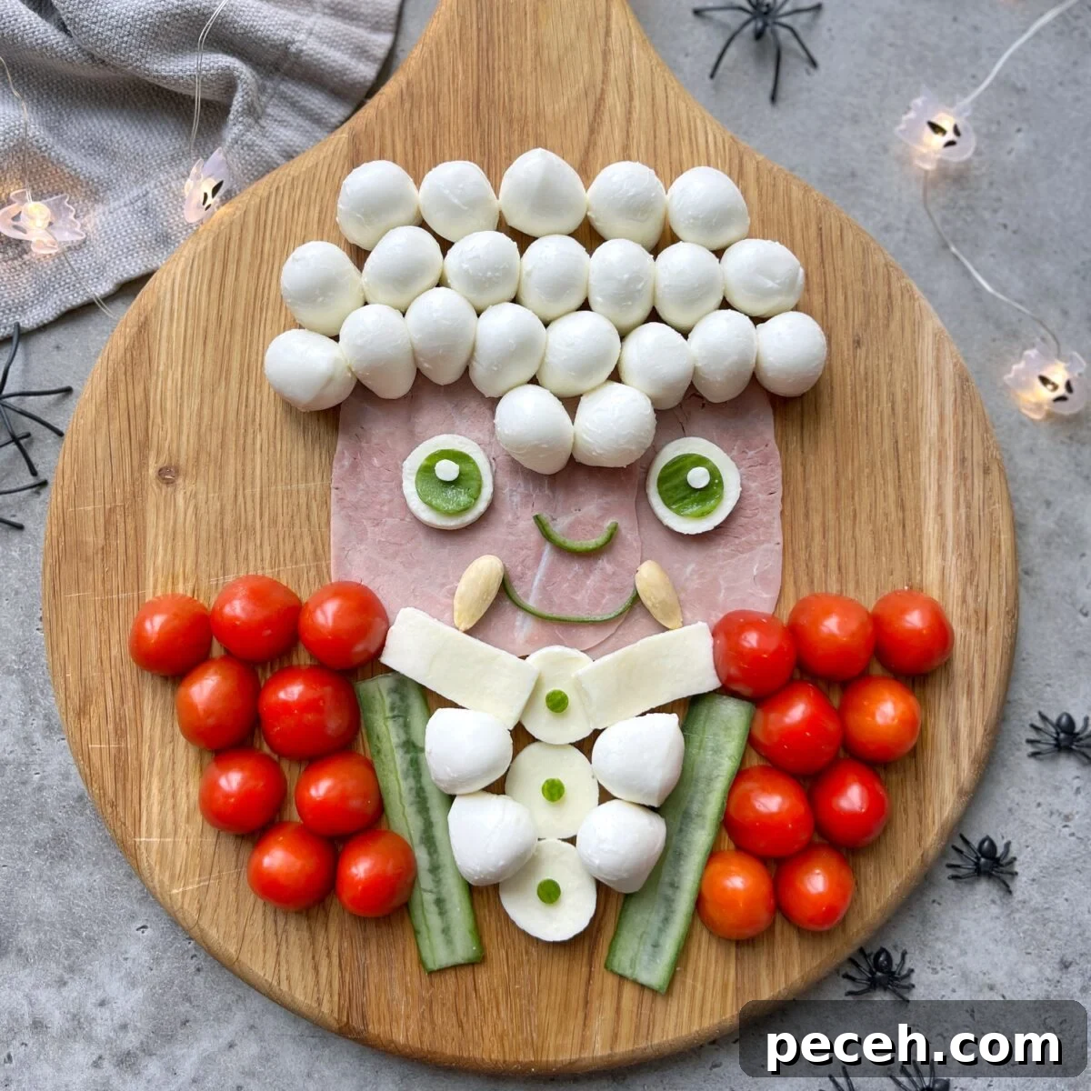 Halloween Charcuterie Board Ideas for a Spooky Party 3 A Vampire face made from ham, mozzarella balls, cucumber, cherry tomatoes, and cheese is arranged on a wooden board. Surrounding it is small plastic spiders and string lights.