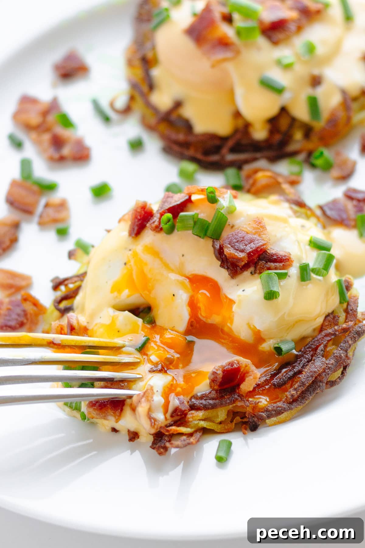Rösti Eggs Benedict with Crispy Potato Cakes 2 Potato rosti eggs Benedict garnished with chopped chives and crispy bacon pieces with a fork cutting into one.