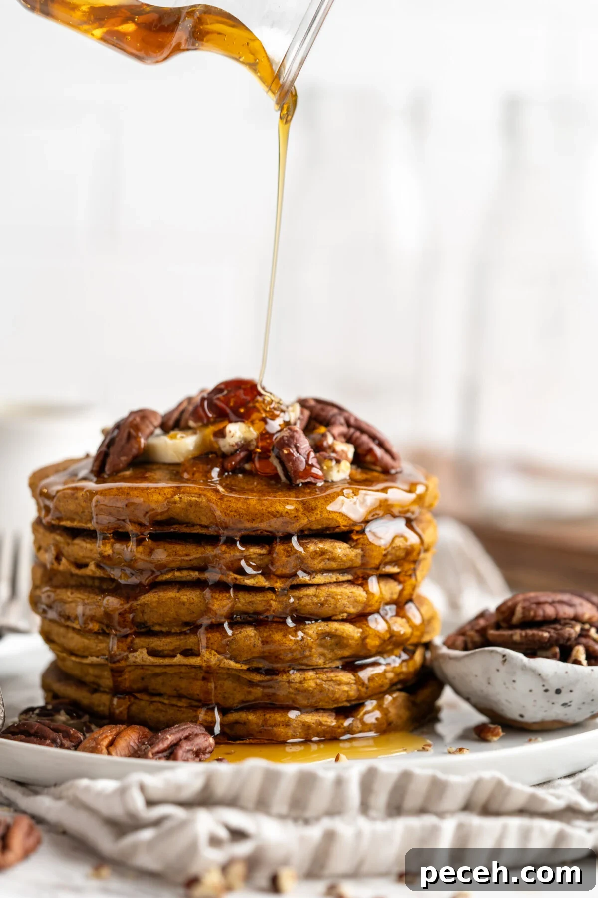 Fluffy vegan pumpkin pancakes are easy to make and loaded with your favorite warm spices. Perfect for a cozy fall breakfast or family brunch! A stack of pumpkin pancakes on a plate with nuts, with syrup being poured over the top.