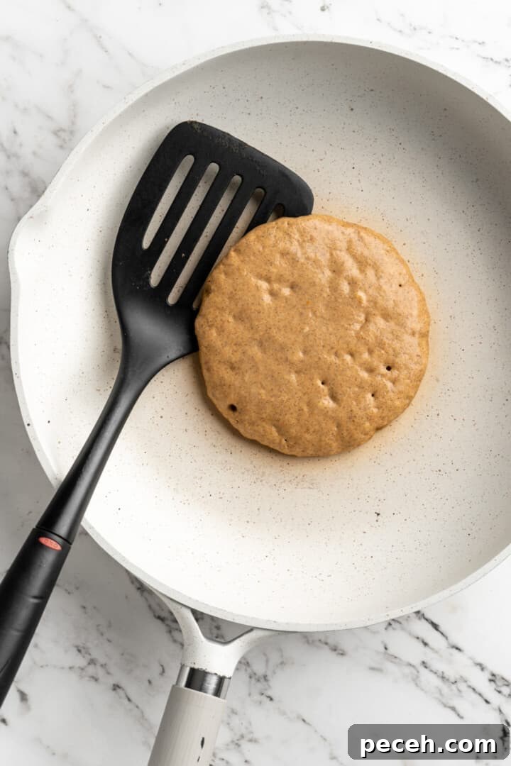 Cook the first side. Overhead view of an uncooked pumpkin pancake in a pan with a spatula.