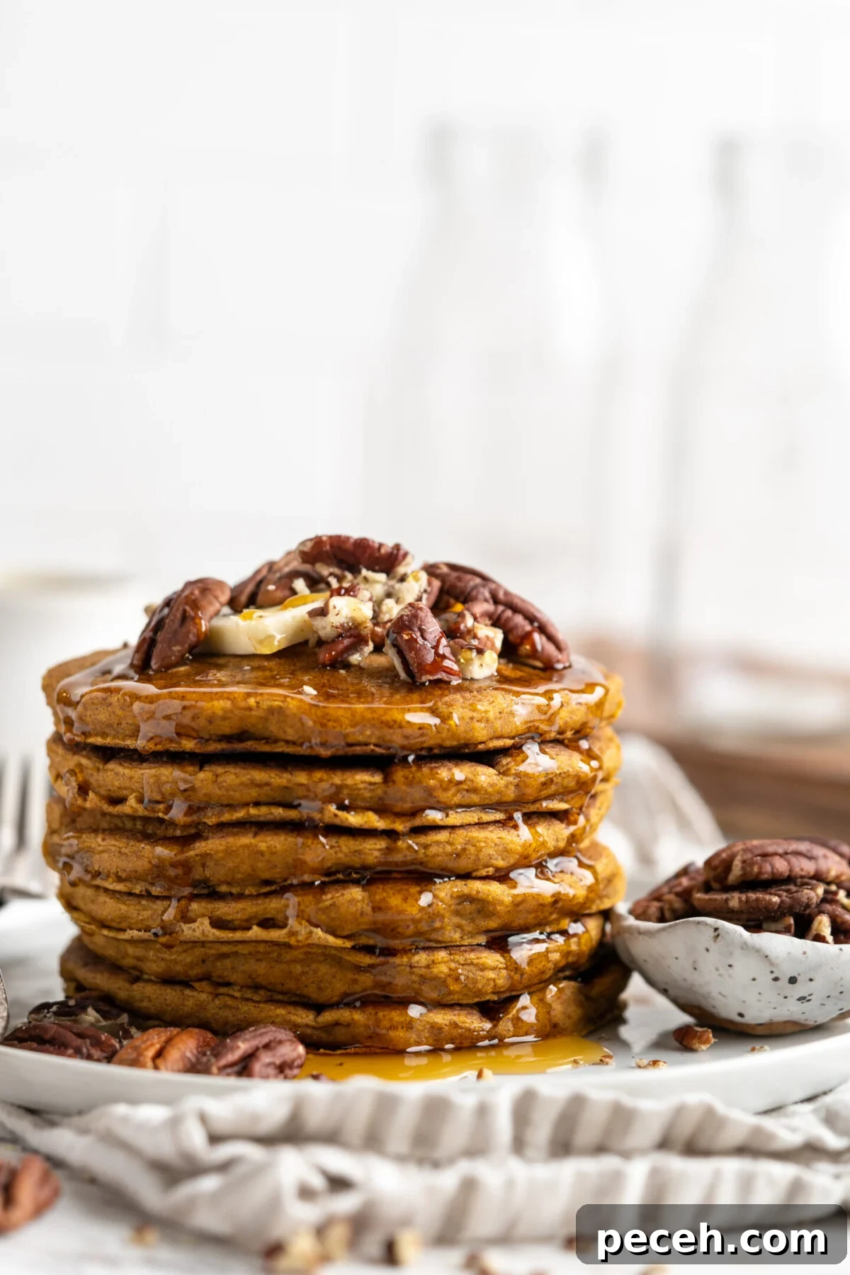 Fluffy vegan pumpkin pancakes are easy to make and loaded with your favorite warm spices. Perfect for a cozy fall breakfast or family brunch! A stack of pumpkin pancakes on a plate, topped with nuts, next to a bowl of nuts.