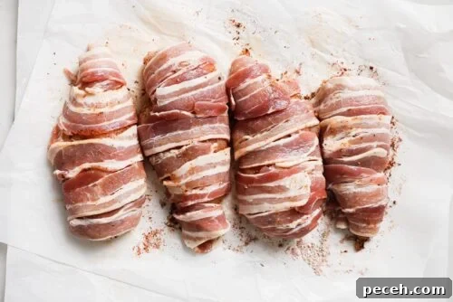 Juicy Bacon-Wrapped Chicken Breast Recipe for Dinner 12 Four Bacon Wrapped Chicken Breasts are arranged on white parchment paper, each wrapped in strips of bacon with a sprinkle of seasoning visible.
