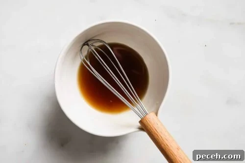 Juicy Bacon-Wrapped Chicken Breast Recipe for Dinner 13 A metal whisk with a wooden handle rests in a white bowl containing a small amount of brown liquid, perhaps the perfect marinade for Bacon Wrapped Chicken Breast, all placed on a light-colored surface.