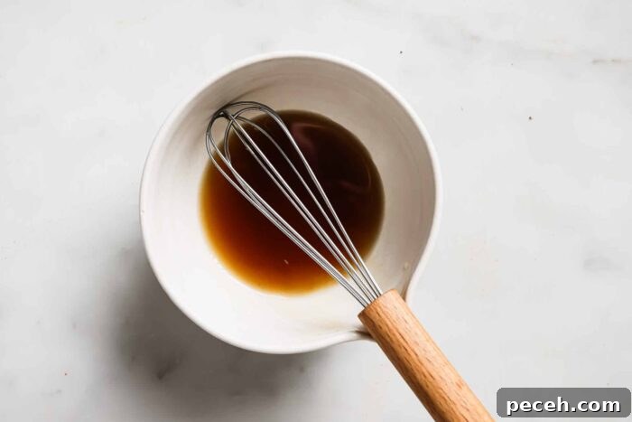 Juicy Bacon-Wrapped Chicken Breast Recipe for Dinner 6 A metal whisk with a wooden handle rests in a white bowl containing a small amount of brown liquid, perhaps the perfect marinade for Bacon Wrapped Chicken Breast, all placed on a light-colored surface.