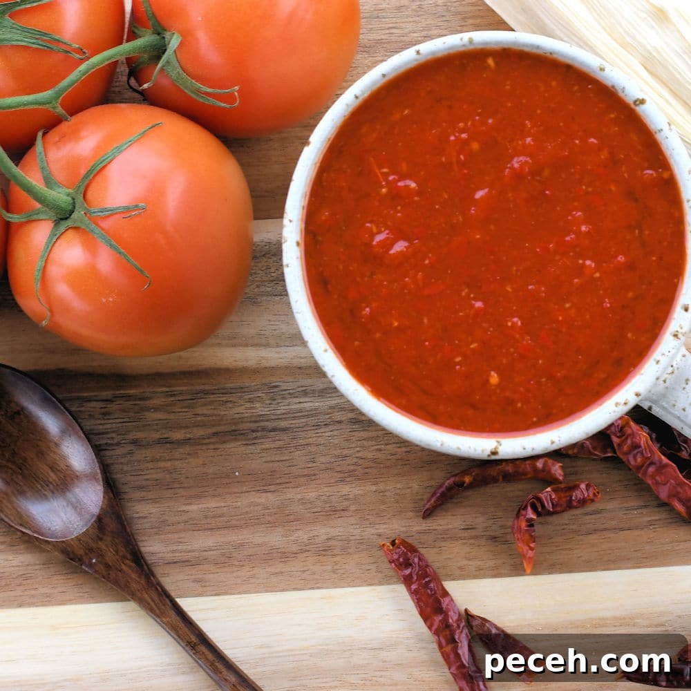 red tamale sauce in a rustic white cup shown on a wooden counter with dried red chiles and tomatoes.