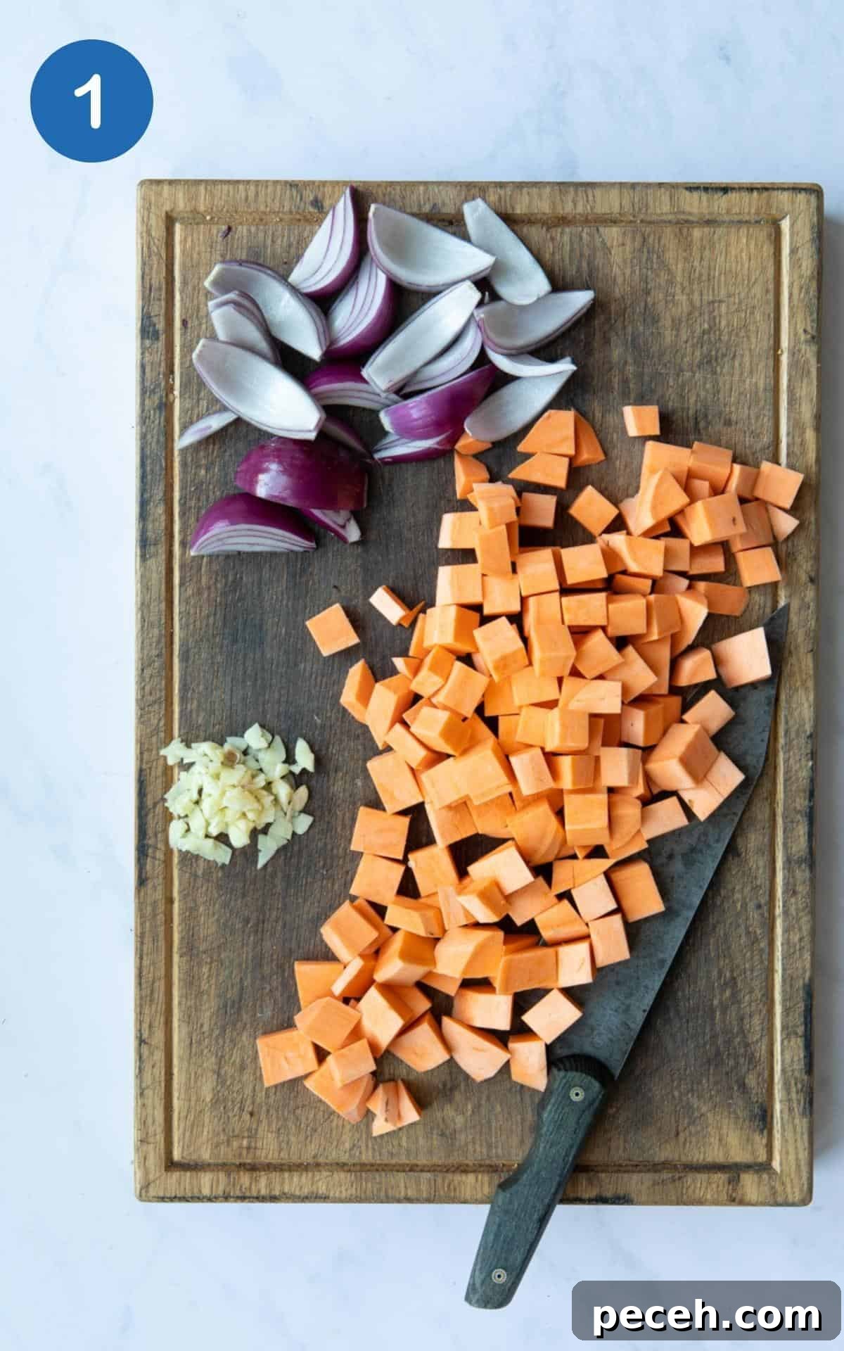 Creamy Sweet Potato Satay Curry with Coconut & Lime 4 chopped sweet potato, finely sliced garlic and onion cut into chunks displayed on a wooden chopping board with knife. Board is displayed on a white marble background.