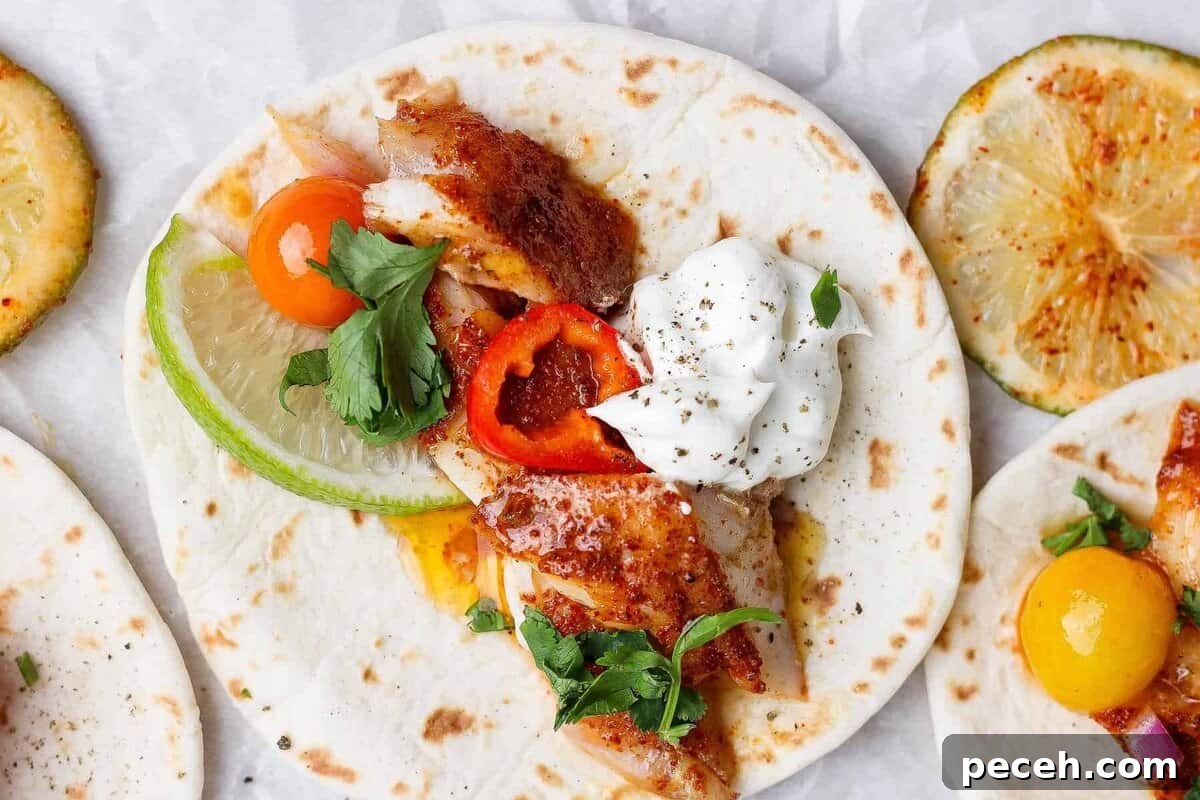 Baked Chile-Lime Fish Tacos with Tangy Cilantro Slaw 2 A soft flour tortilla topped with grilled fish, sliced lime, cherry tomato, red pepper, fresh parsley, and a dollop of sour cream. Grilled lime slices are visible nearby.