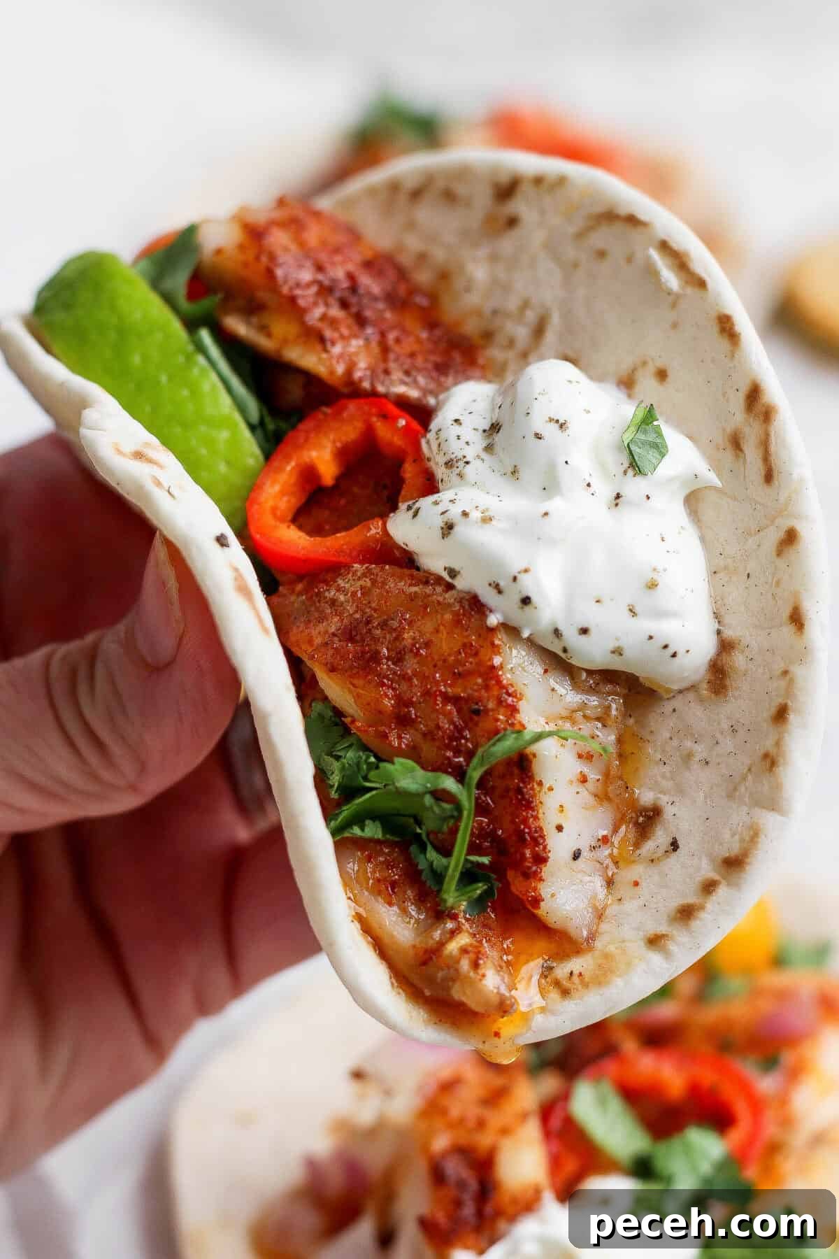 Baked Chile-Lime Fish Tacos with Tangy Cilantro Slaw 5 A hand holds a soft taco filled with seasoned fish, sliced red peppers, fresh greens, avocado, and a dollop of sour cream, garnished with black pepper and herbs.