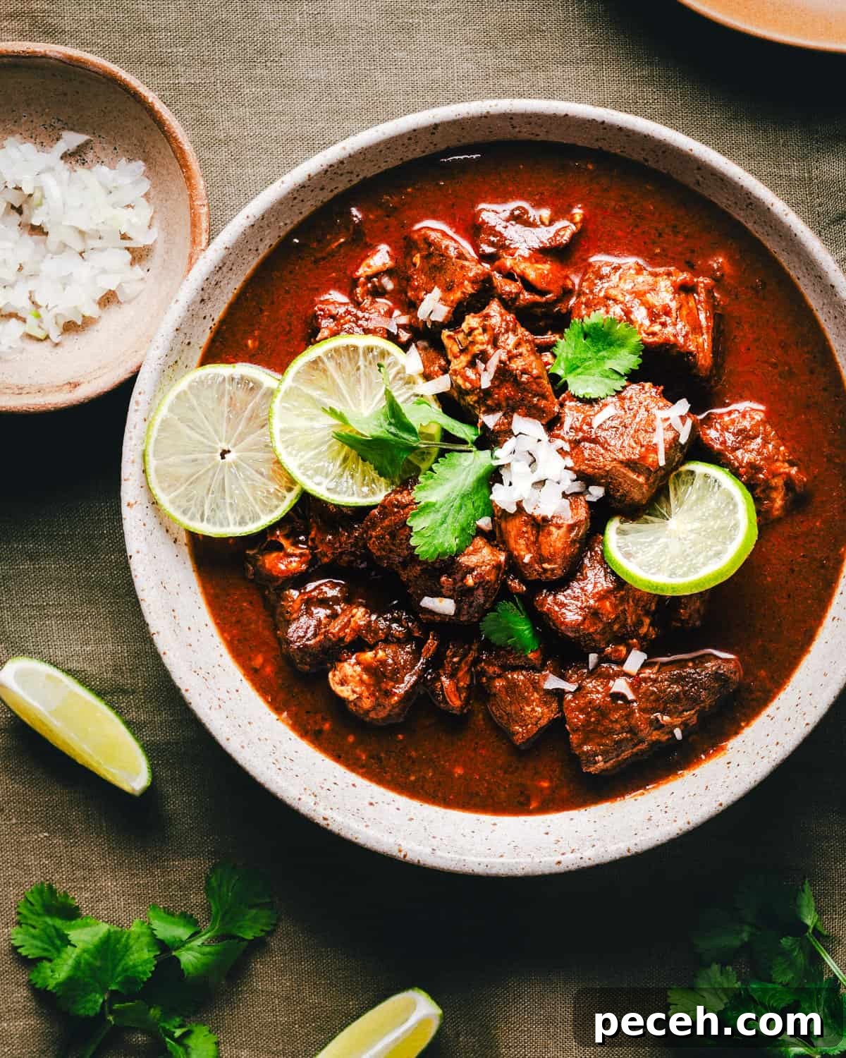 New Mexican Pork Adovada with Red Chile Sauce 2 A bowl of rich Carne Adovada with tender chunks of pork, garnished with sliced limes, cilantro, and diced onions. Set on a dark cloth, the dish is accompanied by a small bowl of onions and lime wedges scattered around.