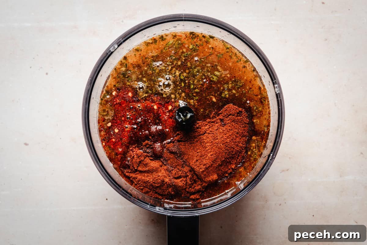 New Mexican Pork Adovada with Red Chile Sauce 9 A food processor contains a blend of spices and liquids, including New Mexico Chile Powder, oil, herbs, and other seasonings, ready to be mixed. The ingredients are vividly colored, with reds and greens visible against the neutral countertop.