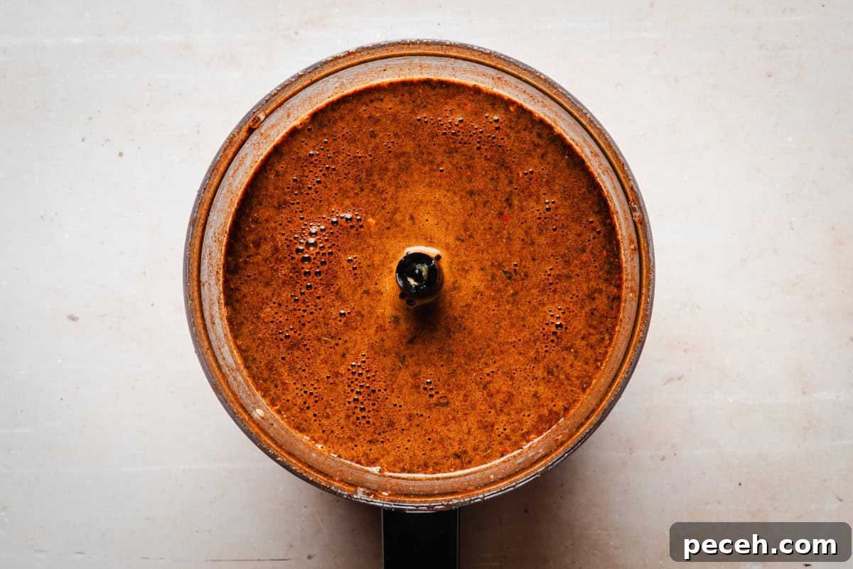 New Mexican Pork Adovada with Red Chile Sauce 10 Top view of a food processor filled with a thick, orange-brown sauce, featuring small bubbles on the surface. The food processor is positioned on a light beige countertop.