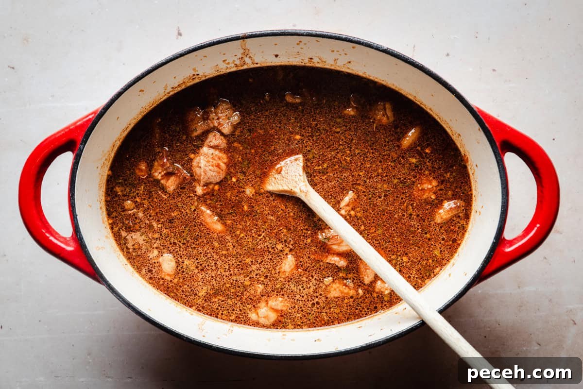 New Mexican Pork Adovada with Red Chile Sauce 11 A red Dutch oven filled with simmering brown stew containing chunks of pork, stirred with a wooden spoon. The stew appears rich and hearty, with visible spices floating on the surface.