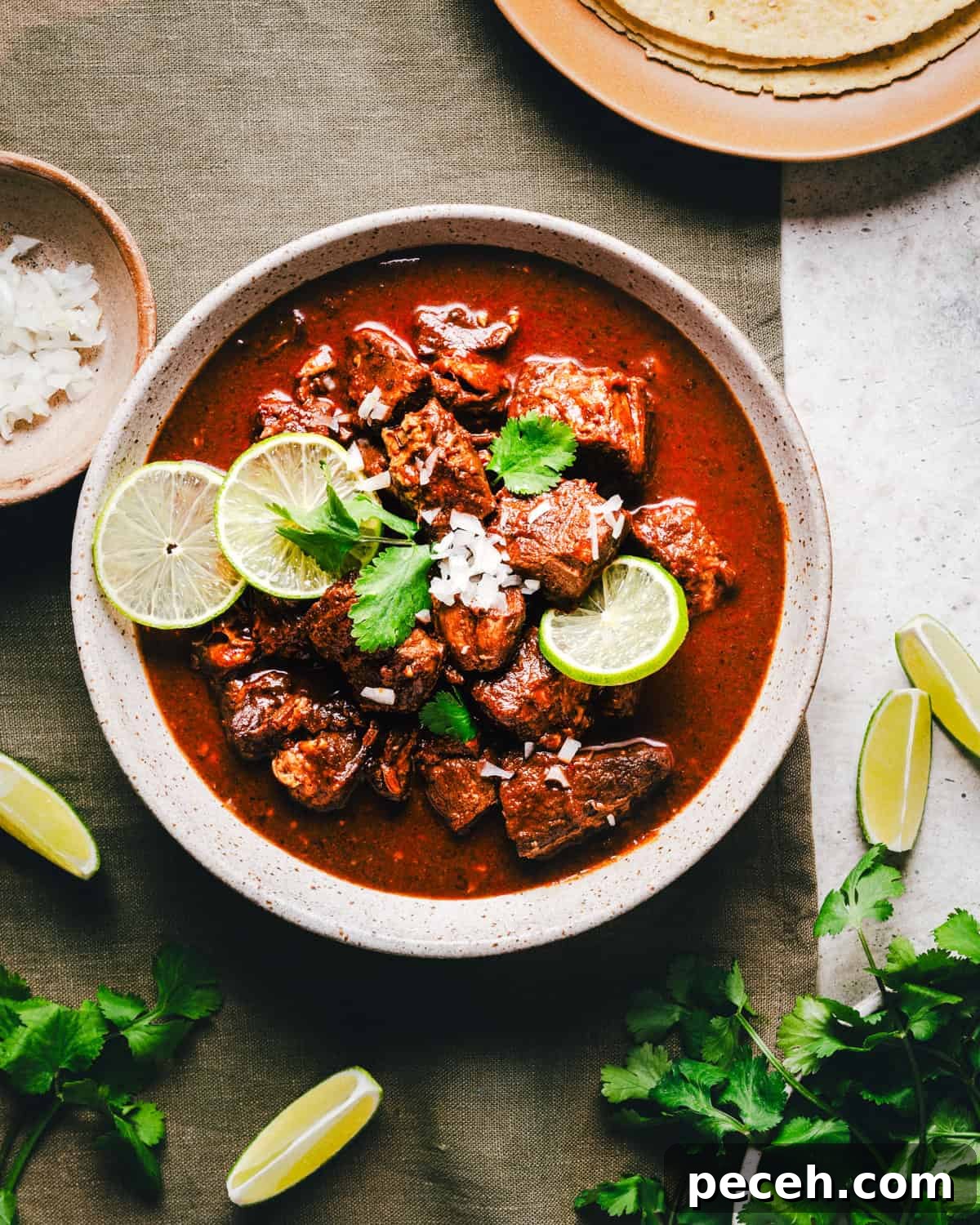 New Mexican Pork Adovada with Red Chile Sauce 12 A bowl of Carne Adovada garnished with lime slices and cilantro sits on a green cloth. Surrounding the bowl are lime wedges, cilantro sprigs, chopped onions in a small bowl, and a plate with tortillas.