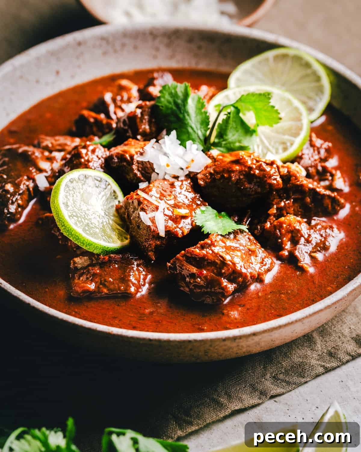 New Mexican Pork Adovada with Red Chile Sauce 13 A bowl of rich Carne Adovada in a red sauce, garnished with sliced lime, fresh cilantro, and a sprinkle of white rice, sits on a textured surface. A small bowl and lime wedges are partially visible in the background.