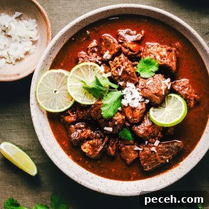 New Mexican Pork Adovada with Red Chile Sauce 19 A bowl of Carne Adovada garnished with lime slices, chopped onions, and cilantro. A small dish with more chopped onions and a lime wedge are on the side, all set on a dark green fabric background.