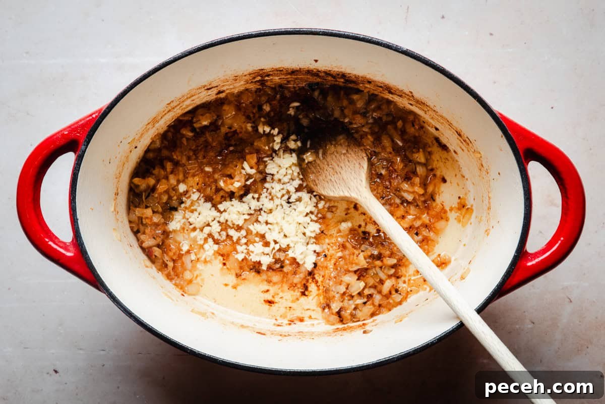 New Mexican Pork Adovada with Red Chile Sauce 8 A red enameled cast iron pot contains sautéed onions with a wooden spoon resting inside. Minced garlic is being added to the mixture, and the pot sits on a speckled light gray surface.